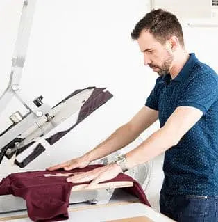 Man using a silkscreen printing press to print on a maroon shirt.